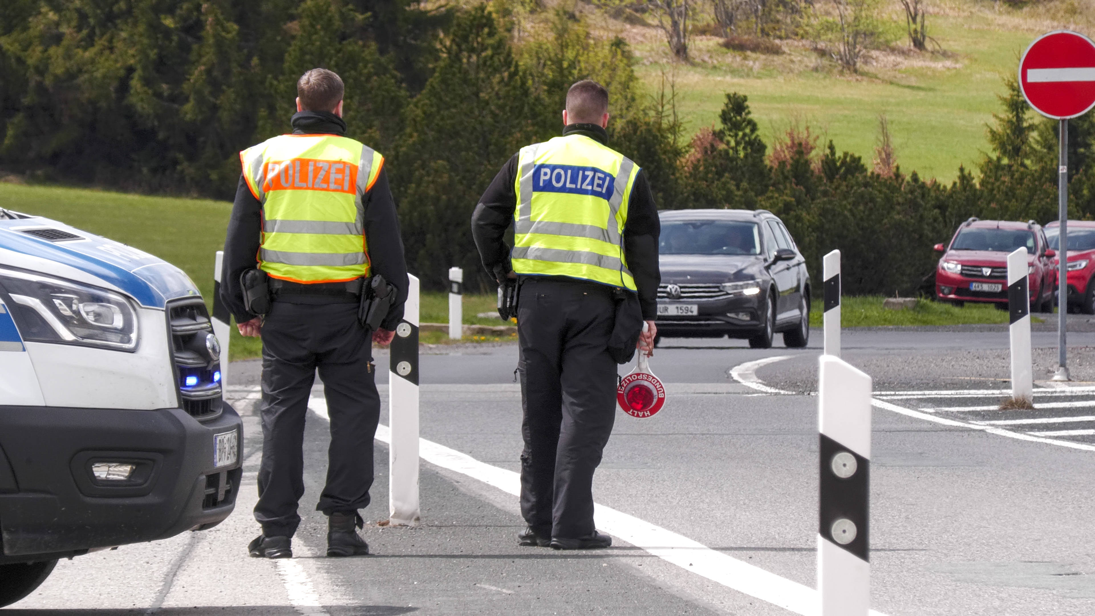 Zwei Männer stehen in gelben Polizeiwesten an einer Bundesstraße. Ein Auto kommt ihnen entgegen. Einer der Männer hält eine Kelle in der Hand.