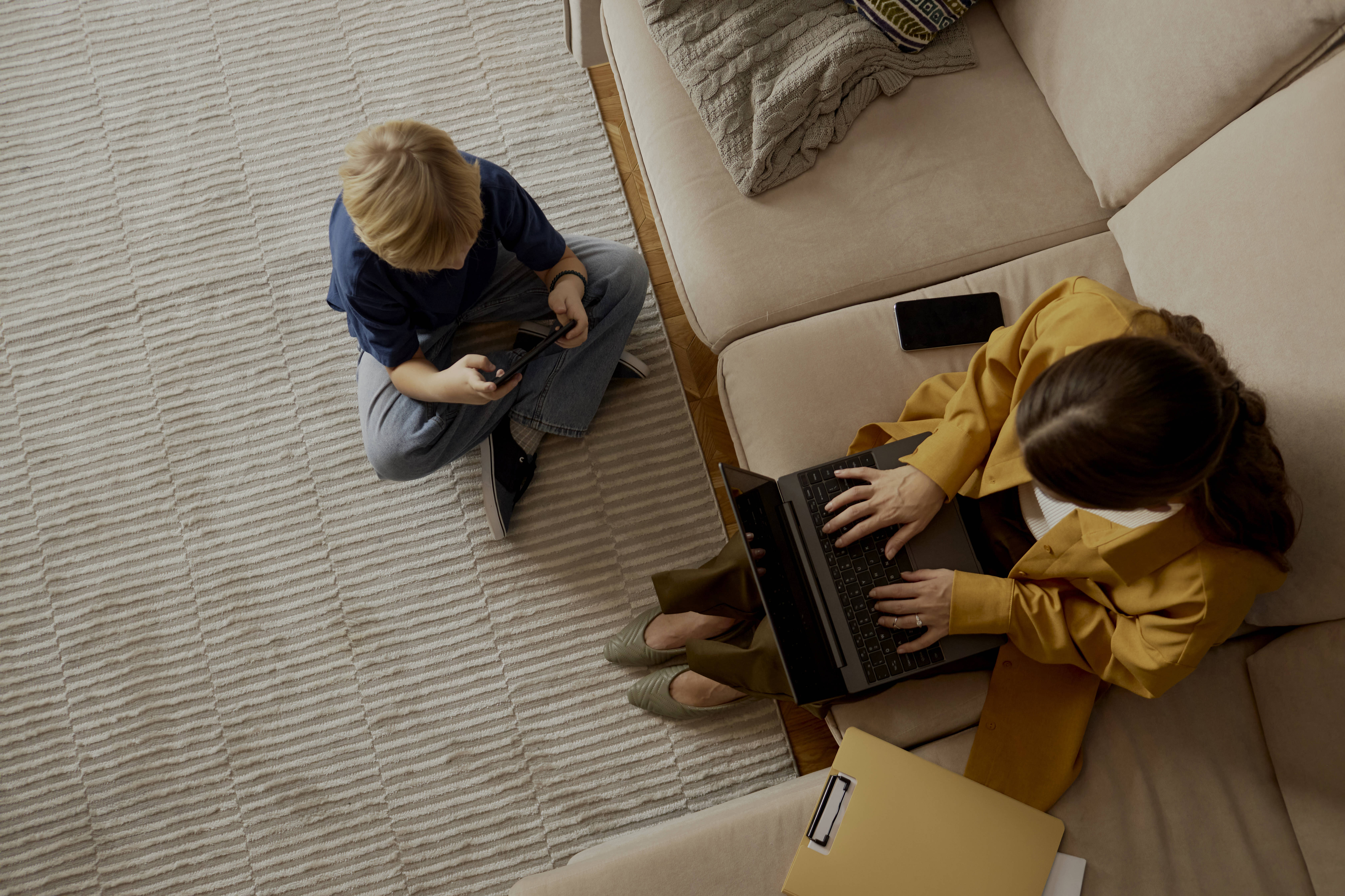 Eine Frau sitzt auf einem Sofa an einem Laptop, daneben ein Kind am Handy.