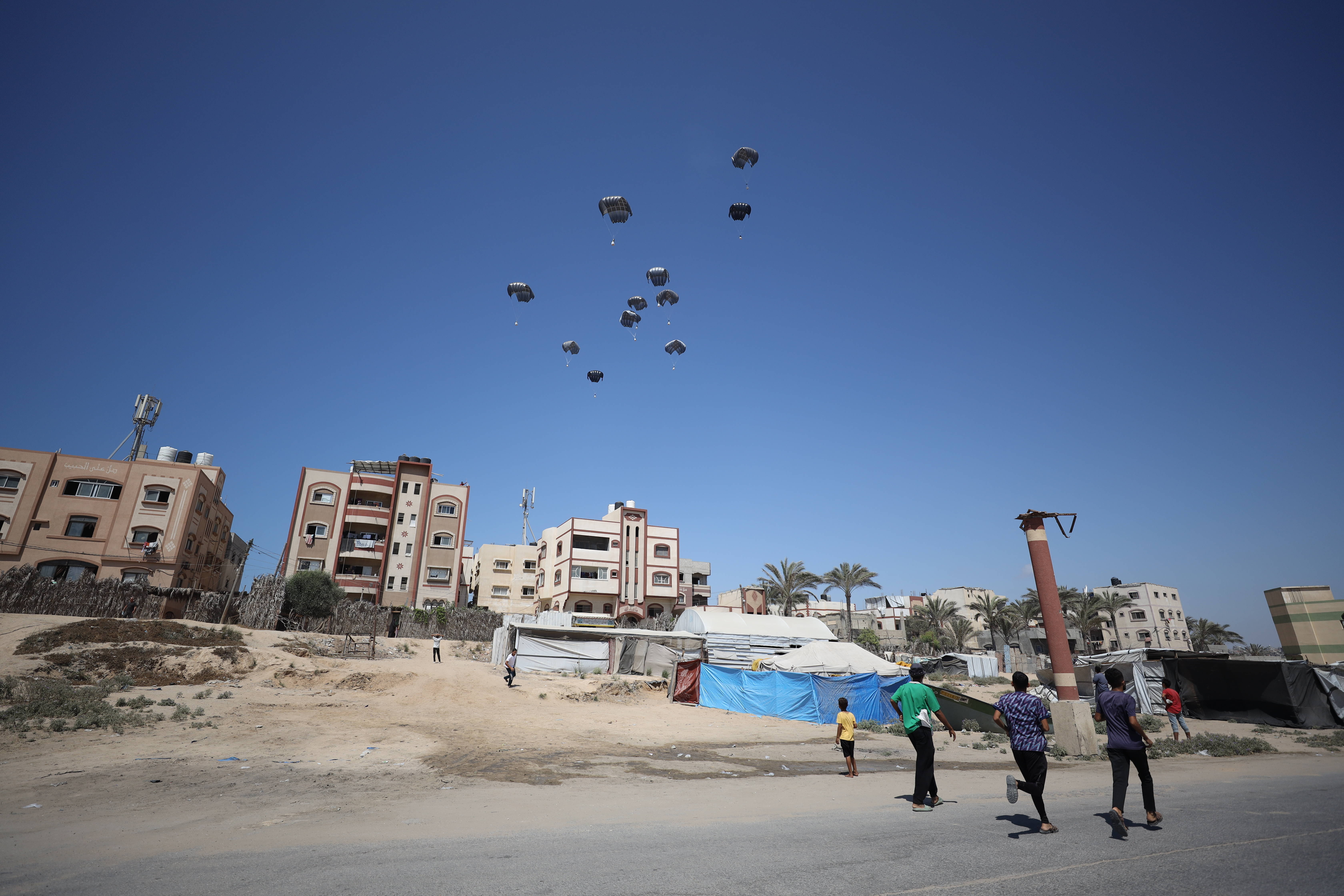 Hilfsgüter an Fallschirmen über Gebäude und Zelten in Gaza, im Vordergrund rechts drei Erwachsene und zwei Kinder