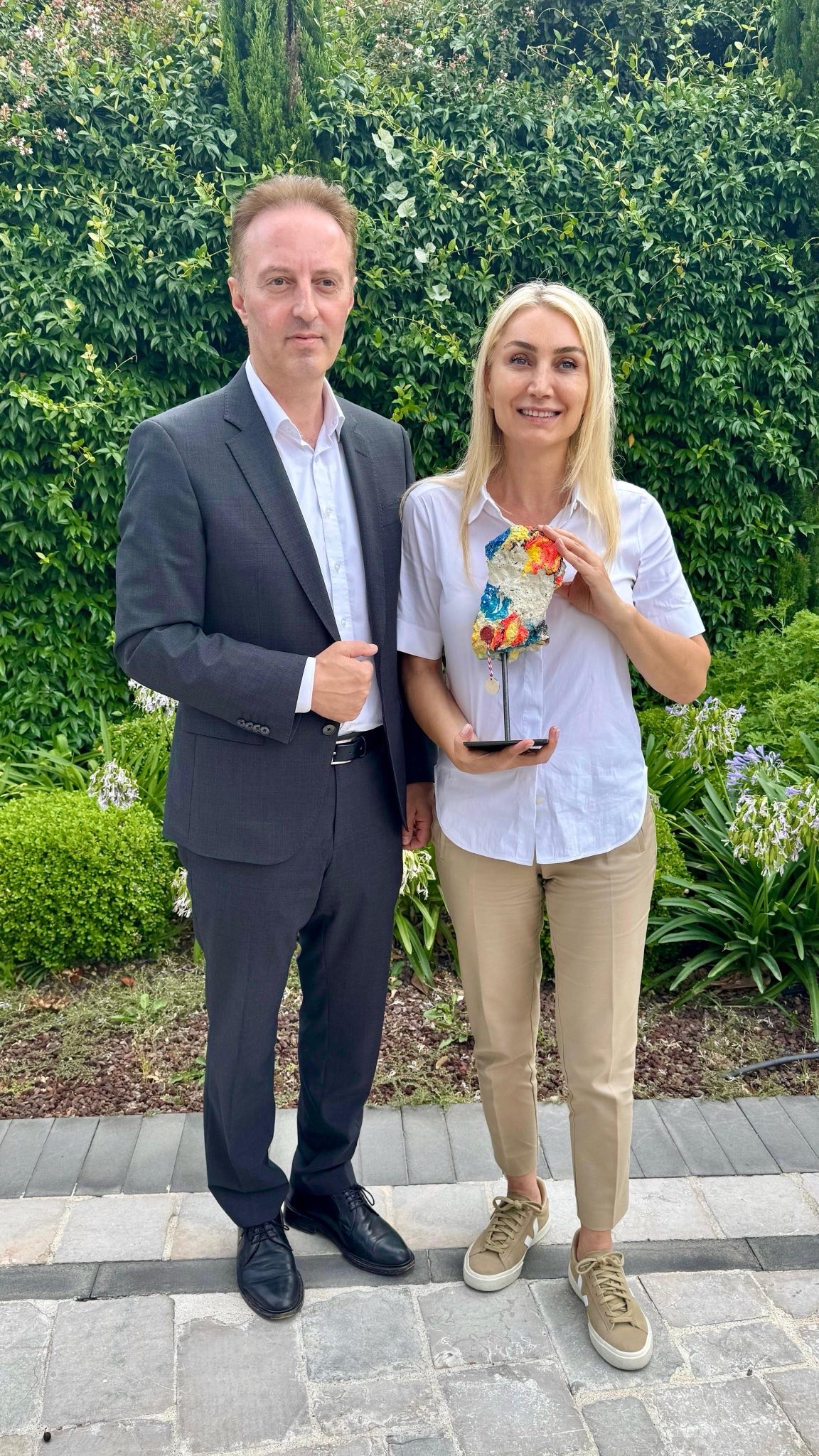 Macit Karahmetoglu und Dilek Imamoglu vor einer grünen Hecke mit einem bemalten Stück Berliner Mauer