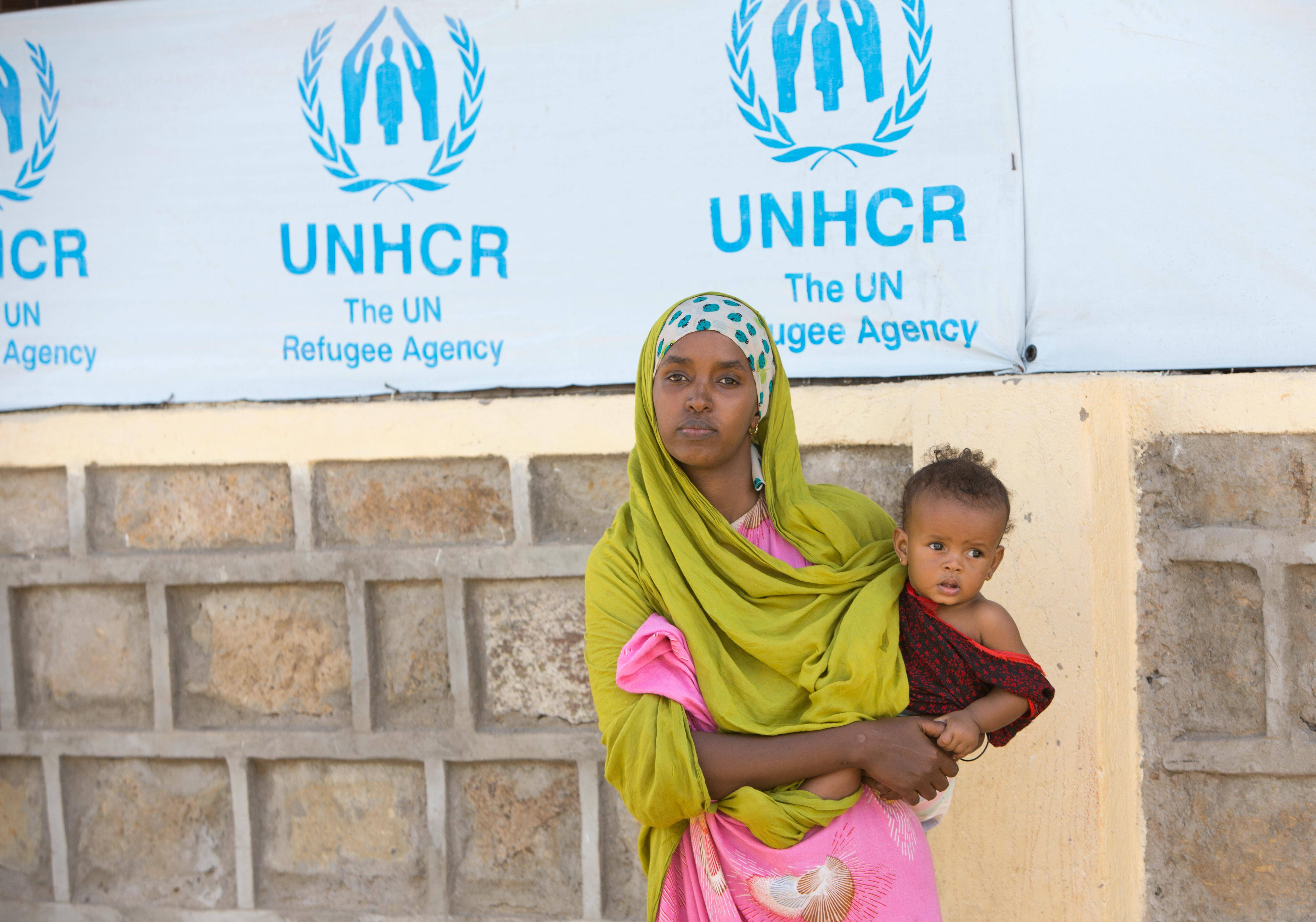 Eine geflüchtete Frau mit ihrem Kind in Kenia vor einem Schild des UN-Flüchtlingshilfswerks.