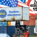 Symbolbild: Ein Gabelstapler mit einen Container auf der Gabel mit der Aufschrift "EU-Handelsabkommen", im Hintergrund eine US-Flagge und ein Warnschild mit der Aufschrift "Zoll 15 Prozent"