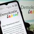 Internetseite von "Demokratie leben!" auf einem Smartphone, im Hintergrund leicht unscharf die Seite auf einem Monitor