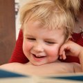 Ein lachendes Kind auf dem Schoß eines Erwachsenen, im Vordergrund im Anschnitt ein Buch
